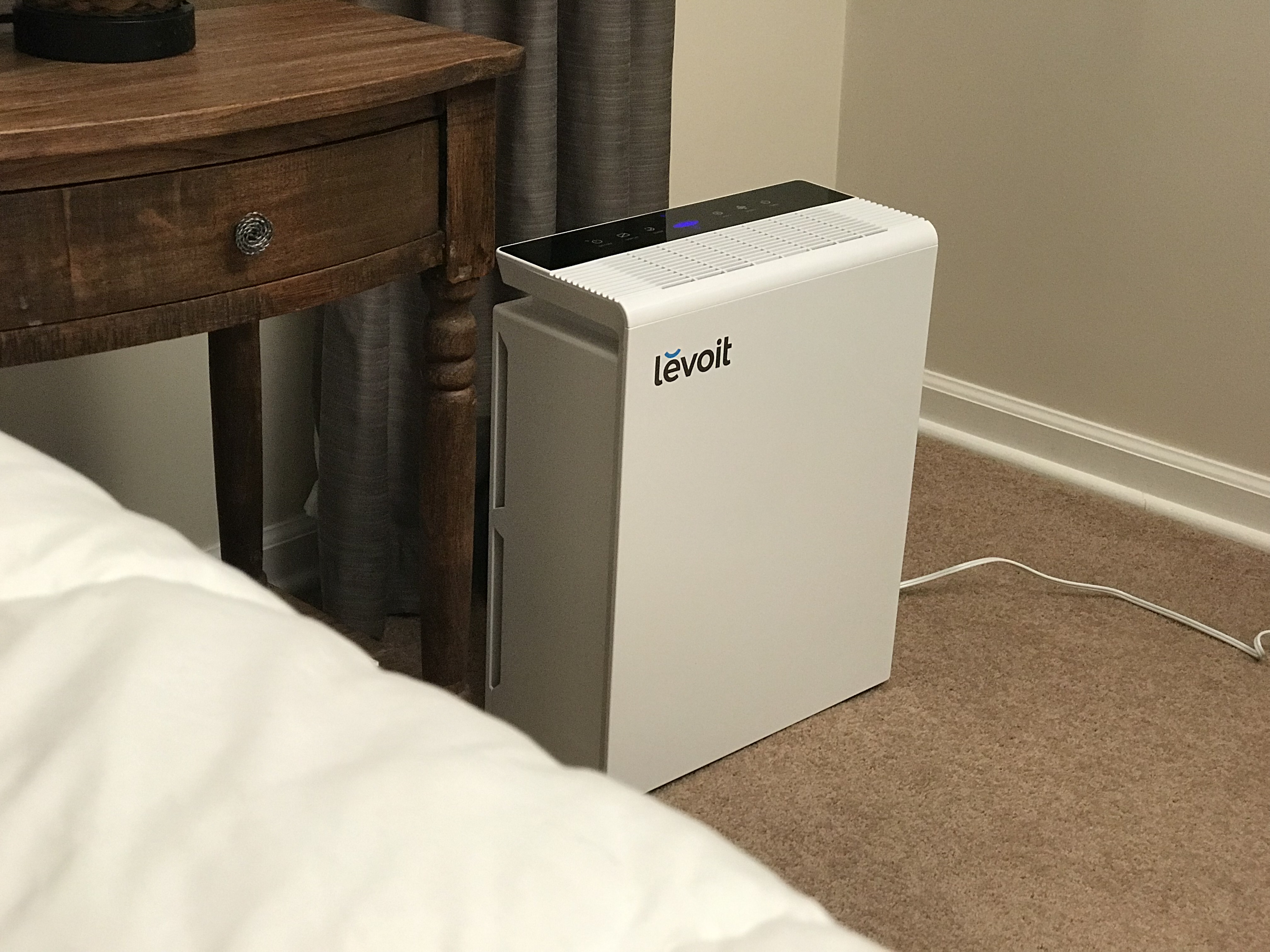 Keeping an air purifier running in your bedroom while you sleep is a great habit to start so you can sleep better and breathe cleaner air. (Alt: White Levoit air purifier running beside bedroom nightstand at night)
”Air Purifier (Levoit LV-PUR131S) (49318413416)” by Home Air Quality Guides is licensed under CC BY-SA 2.0.